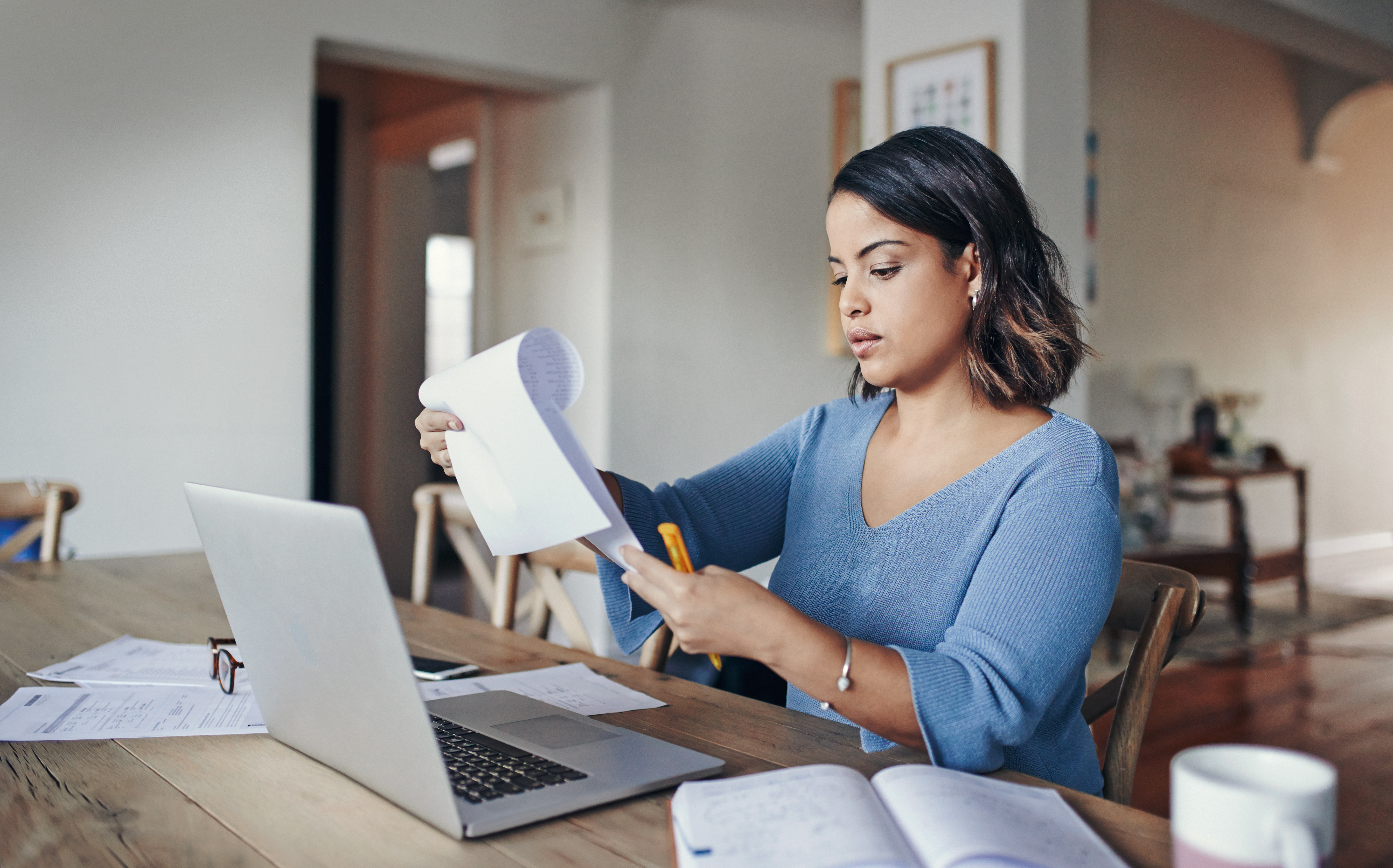 Een persoon die vanuit huis werkt, kijkt naar een papieren document en zit aan een tafel met een laptop.